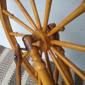 May include: A wooden spinning wheel featuring a large circular frame with multiple spokes radiating from a central hub. The structure is crafted from light brown wood with a smooth finish, showing a vertical support post and a small metal component.