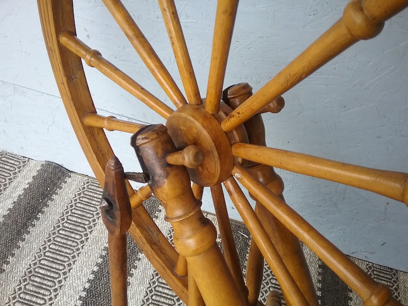 May include: A wooden spinning wheel featuring a large circular frame with multiple spokes radiating from a central hub. The structure is crafted from light brown wood with a smooth finish, showing a vertical support post and a small metal component.