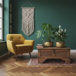 Può includere: Una poltrona imbottita color giallo senape con bottoni e gambe in legno scuro, posizionata accanto a un baule in legno intagliato con due fioriere in ottone contenenti una monstera e uno spatifillo, e un arazzo in macramè appeso sopra.