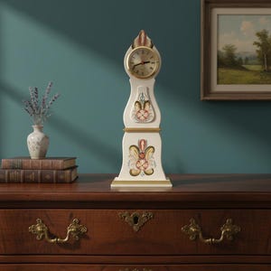 May include: A cream-coloured, decorative longcase clock with a gold clock face and floral patterns. The clock sits on a dark wood chest of drawers with brass handles. A vase of lavender and a framed painting are in the background.