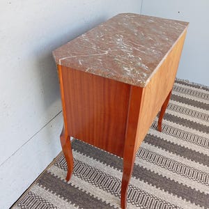 May include: A vintage wooden cabinet with a marble top. The cabinet has a reddish-brown finish and curved legs. The marble top features a reddish-brown and white veined pattern. The cabinet is positioned on a patterned rug.