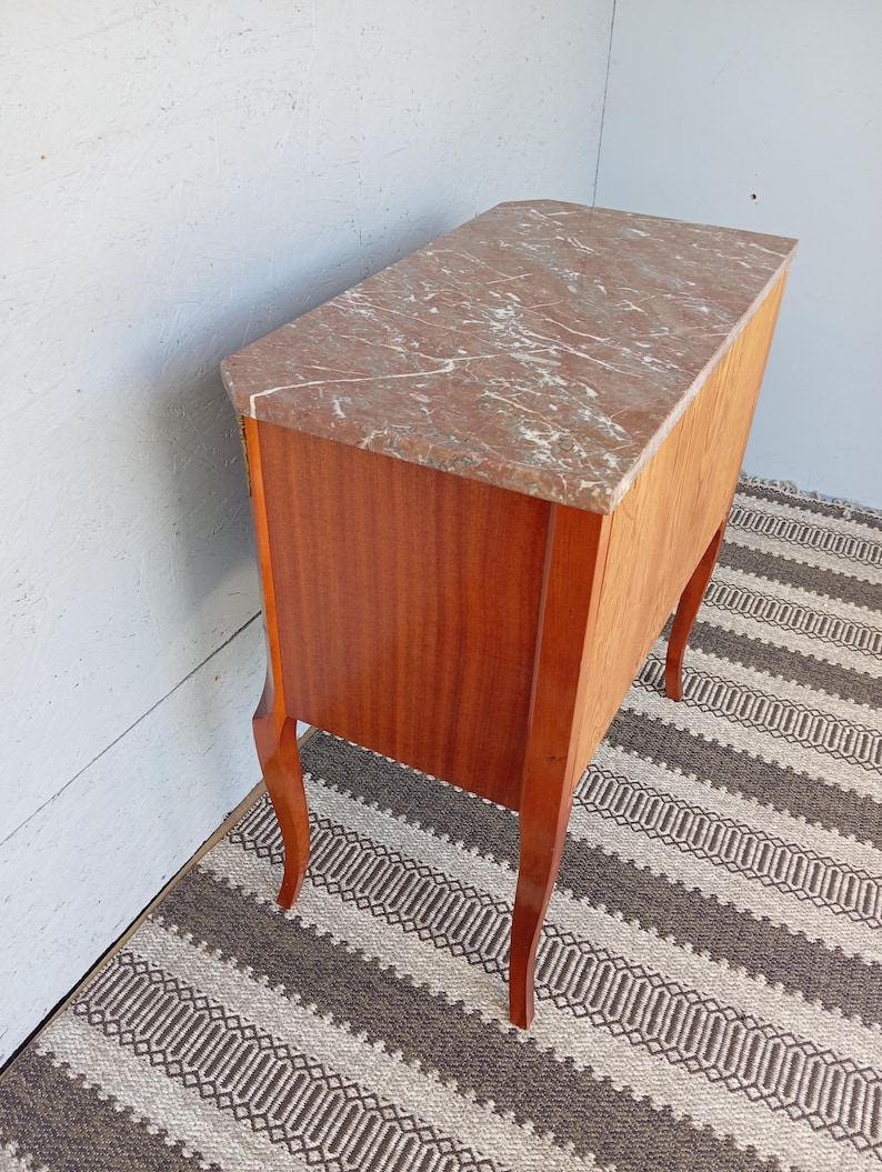 May include: A vintage wooden cabinet with a marble top. The cabinet has a reddish-brown finish and curved legs. The marble top features a reddish-brown and white veined pattern. The cabinet is positioned on a patterned rug.