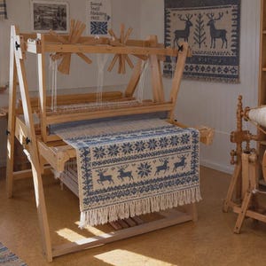 May include: A wooden floor loom with a partially woven textile featuring a blue and white pattern of reindeer and snowflakes. The fabric has a fringed edge and is held within the frame of the loom, which includes wooden spools and white thread.