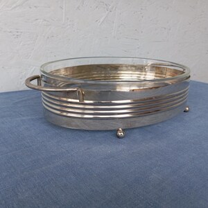 May include: A silver serving dish with a glass insert and a decorative, ribbed design. The dish has two handles and rests on four small feet.