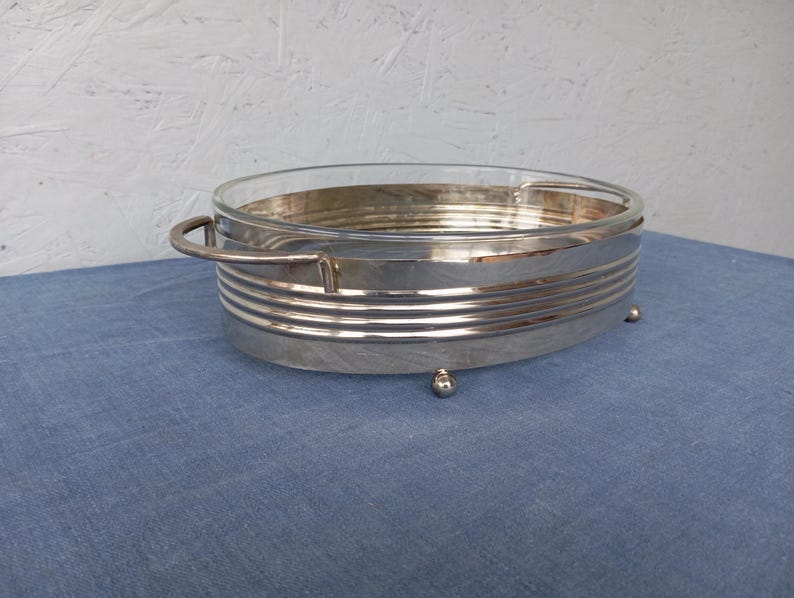 May include: A silver serving dish with a glass insert and a decorative, ribbed design. The dish has two handles and rests on four small feet.
