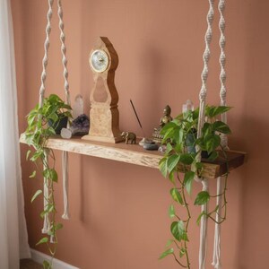 May include: A natural wood floating shelf suspended by two thick, cream-colored macrame ropes with twisted patterns. The shelf holds a wooden clock, two potted green pothos plants with trailing vines, crystal clusters, a small elephant figurine, and incense.
