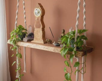 May include: A natural wood floating shelf suspended by two thick, cream-coloured macrame ropes with twisted patterns. The shelf holds a wooden clock, two potted green pothos plants with trailing vines, crystal clusters, a small elephant figurine, and incense.