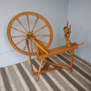 May include: A light brown wooden spinning wheel featuring a large circular wheel with multiple spokes, a horizontal frame, and a vertical flyer assembly. The structure is supported by three legs and includes a treadle mechanism for manual operation.