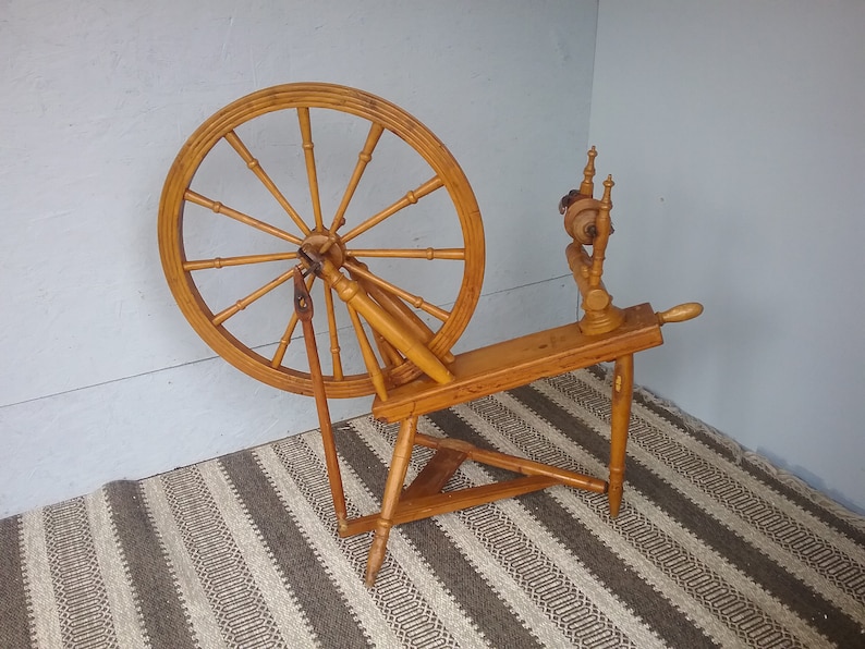 May include: A light brown wooden spinning wheel featuring a large circular wheel with multiple spokes, a horizontal frame, and a vertical flyer assembly. The structure is supported by three legs and includes a treadle mechanism for manual operation.