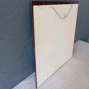May include: A square wall plaque featuring a plain white back panel with a brown plastic frame edge. A thin, light-colored twisted cord is attached to the top of the frame for hanging purposes. The surface of the white panel shows minor marks.