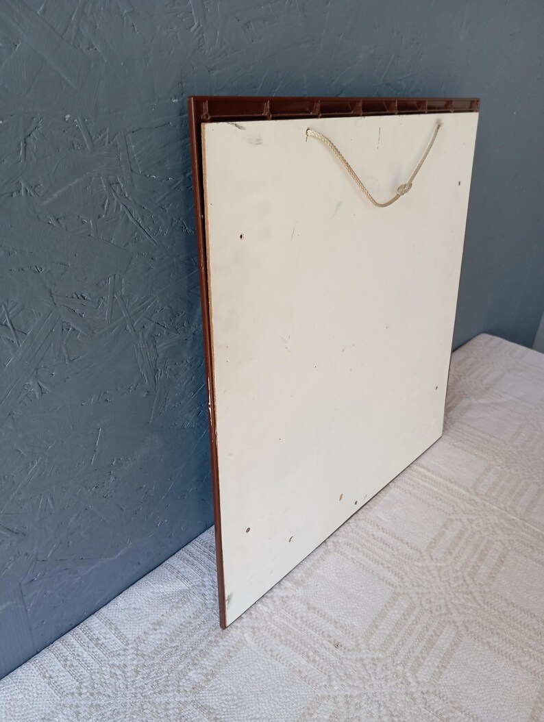 May include: A square wall plaque featuring a plain white back panel with a brown plastic frame edge. A thin, light-colored twisted cord is attached to the top of the frame for hanging purposes. The surface of the white panel shows minor marks.
