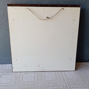 May include: A square wall-mounted display board featuring a smooth white front panel with a dark brown frame. A thin, light-colored string is attached to the top for hanging purposes. The surface shows four small circular holes near the corners.