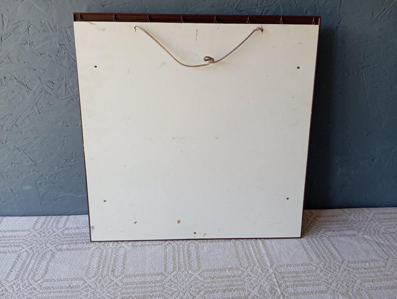 May include: A square wall-mounted display board featuring a smooth white front panel with a dark brown frame. A thin, light-colored string is attached to the top for hanging purposes. The surface shows four small circular holes near the corners.