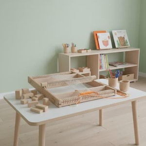 May include: A wooden weaving loom with white yarn and orange thread sits on a white table. Wooden blocks are scattered nearby. A bookshelf in the background holds books and art supplies. The room has a light green wall.