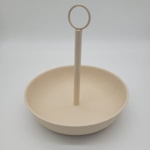 Könnte beinhalten: Eine weiße Keramikschüssel mit einem Metallgriff. Die Schüssel ist rund und hat eine strukturierte Oberfläche. Der Griff ist in der Mitte der Schüssel befestigt und hat oben einen Ring.