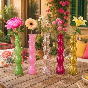 Könnte beinhalten: Fünf farbige Glasvasen in Grün, Rosa, Transparent, Magenta und Gelb, jede mit einer anderen Blume. Die Blumen umfassen eine rosa Pfingstrose, eine pfirsichfarbene Gerbera und weiße Gänseblümchen. Auf einem Holztisch.