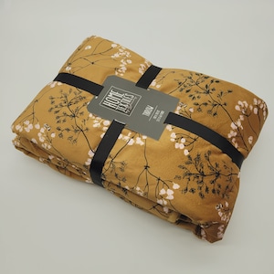 Könnte beinhalten: Eine senfgelbe Kuscheldecke mit einem Blumenmuster aus kleinen weißen Blüten und schwarzen Stielen. Die Decke ist gefaltet und mit einem schwarzen Band umwickelt. Das Etikett auf dem Band lautet "Home Textiles by Designer's Choice" und "Throw 50" x 60" 100% Polyester."