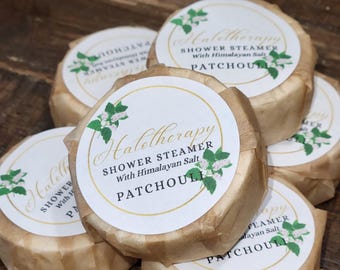 May include: A stack of six shower steamers wrapped in brown paper. Each steamer has a white label with the text "Halotherapy Shower Steamer With Himalayan Salt Patchouli" and a floral design. The background is a dark wood surface.