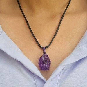 Könnte beinhalten: Eine Halskette mit einem Anhänger aus lila Amethyst und einem schwarzen Band. Der Amethyst ist mit lila Draht umwickelt. Die Halskette wird über einem hellblauen Poloshirt getragen.
