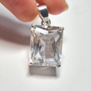 Puede incluir: Un colgante rectangular de cristal transparente con una fianza plateada es sostenido por una mano sobre un fondo blanco. El cristal está facetado, captando la luz y creando un efecto brillante. El colgante es una pieza de joyería clásica.