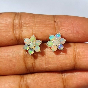Puede incluir: Un par de pendientes de botón con forma de flor. Cada pendiente presenta siete piedras preciosas de ópalo redondas e iridiscentes engastadas en un metal color plata. Los ópalos muestran una gama de colores, incluyendo blanco, verde y azul.