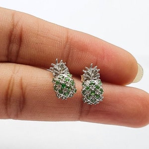 Puede incluir: Un par de pendientes de piña de plata con piedras preciosas verdes. Los pendientes son pequeños y delicados, y serían un gran regalo para alguien que ama las piñas.