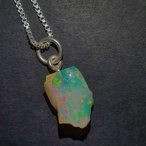 Op de afbeelding: Een zilveren ketting met een ruwe opaal hanger. De opaal toont een levendig kleurenspel met flitsen van groen, roze en geel. De hanger is bevestigd aan een zilveren ketting, wat een uniek en opvallend sieraad creëert.