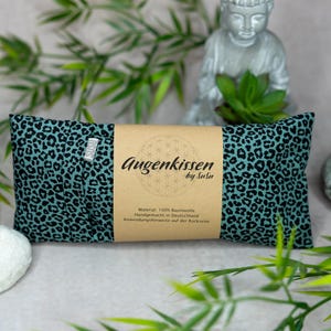 Könnte beinhalten: Ein rechteckiges Augenkissen mit türkisem Leopardenmuster. Das Kissen ist mit einem braunen Papieretikett mit Text umwickelt. Eine kleine Statue und etwas Grün befinden sich im Hintergrund.