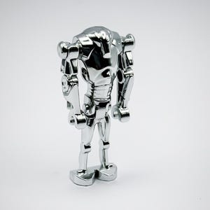Puede incluir: Figura de robot cromado con un diseño detallado. El robot tiene una forma humanoide con extremidades articuladas y una cabeza redondeada. La superficie metálica refleja la luz, resaltando las características mecánicas. La figura se apoya sobre una base rectangular.
