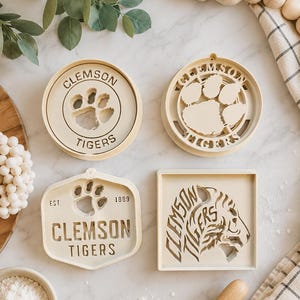 May include: Four off-white plastic cookie cutters featuring Clemson Tigers branding. Two are circular with paw prints, one is a hexagon with a paw print and the text EST. 1889, and one is a square featuring a tiger head profile and Clemson Tigers text.