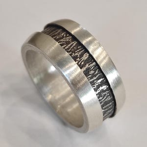 Op de afbeelding: Een brede ring met een gepolijste zilveren afwerking aan de buitenranden en een verzonken middenkanaal met een donker, geoxideerd en sterk getextureerd oppervlak dat lijkt op ruwe boomschors of organische geologische formaties.
