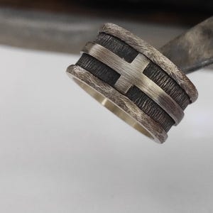 Puede incluir: Un anillo de banda ancha con un centro texturizado oscuro y oxidado, superpuesto con una cruz de plata pulida. Los bordes del anillo tienen un acabado rugoso y desgastado que contrasta con el interior liso y brillante de plata.