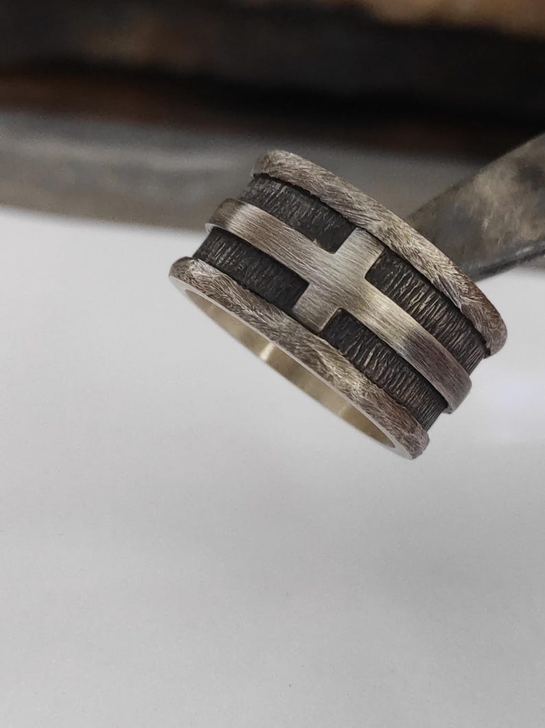 Puede incluir: Un anillo de banda ancha con un centro texturizado oscuro y oxidado, superpuesto con una cruz de plata pulida. Los bordes del anillo tienen un acabado rugoso y desgastado que contrasta con el interior liso y brillante de plata.