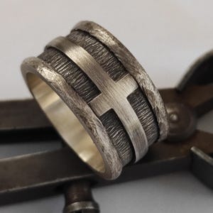 Puede incluir: Un anillo ancho de plata con una banda central texturizada y oxidada oscura, con un motivo de cruz de plata liso y elevado. Los bordes del anillo tienen un acabado rugoso y martillado, y la superficie interior es lisa y pulida.