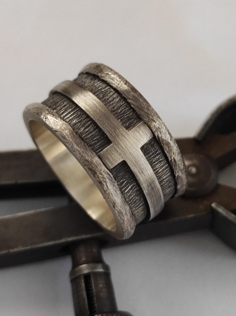 Puede incluir: Un anillo ancho de plata con una banda central texturizada y oxidada oscura, con un motivo de cruz de plata liso y elevado. Los bordes del anillo tienen un acabado rugoso y martillado, y la superficie interior es lisa y pulida.