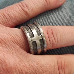 Puede incluir: Un anillo ancho de plata que presenta un emblema de cruz pulida en el centro sobre un fondo oscuro, texturizado y hundido. La banda tiene bordes elevados de plata cepillada que enmarcan el dise&ntilde;o central, creando un contraste marcado entre la cruz lisa y la superficie met&aacute;lica rugosa y oscurecida.