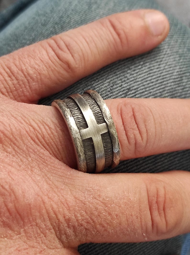 Puede incluir: Un anillo ancho de plata que presenta un emblema de cruz pulida en el centro sobre un fondo oscuro, texturizado y hundido. La banda tiene bordes elevados de plata cepillada que enmarcan el dise&ntilde;o central, creando un contraste marcado entre la cruz lisa y la superficie met&aacute;lica rugosa y oscurecida.