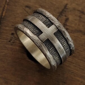 Puede incluir: Un anillo ancho de plata con una banda central texturizada y oxidada oscura, que presenta un emblema de cruz de plata lisa y pulida. Los bordes del anillo tienen un acabado martillado rugoso y el interior es de plata lisa y pulida.