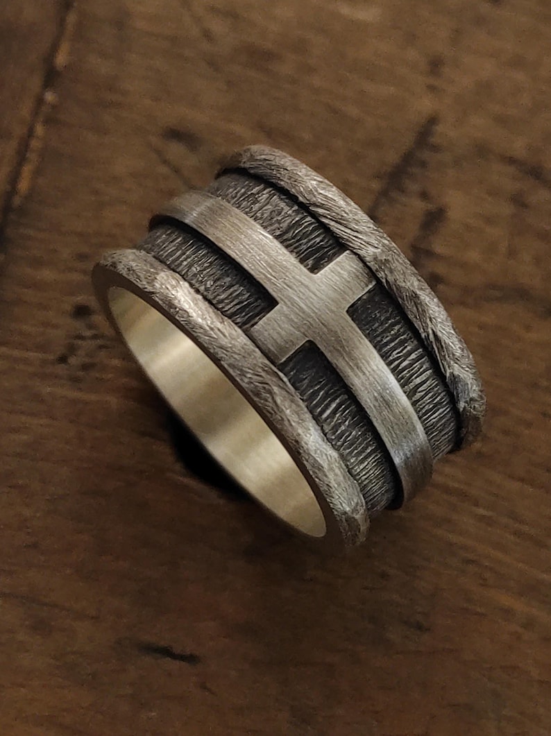 Puede incluir: Un anillo ancho de plata con una banda central texturizada y oxidada oscura, que presenta un emblema de cruz de plata lisa y pulida. Los bordes del anillo tienen un acabado martillado rugoso y el interior es de plata lisa y pulida.