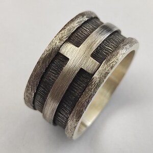 Puede incluir: Un anillo ancho de plata con una banda central oscura y texturizada que presenta un motivo de cruz en plata pulida en relieve. Los bordes del anillo tienen un acabado martillado rugoso que contrasta con el brillo met&aacute;lico liso de la cruz.