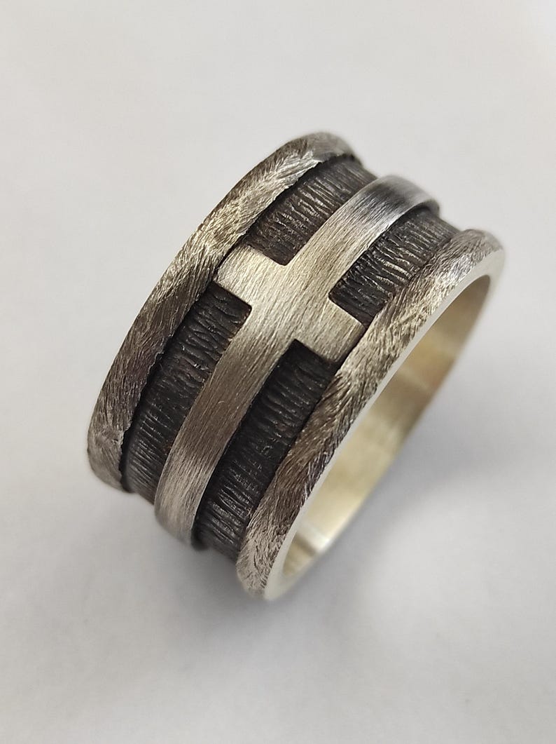 Puede incluir: Un anillo ancho de plata con una banda central oscura y texturizada que presenta un motivo de cruz en plata pulida en relieve. Los bordes del anillo tienen un acabado martillado rugoso que contrasta con el brillo met&aacute;lico liso de la cruz.