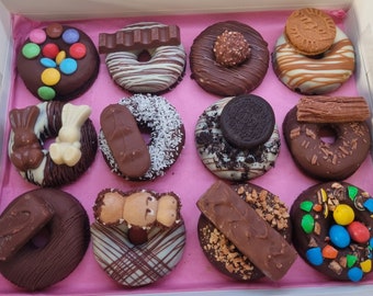 Op de afbeelding: Een doos met twaalf chocolade donuts, elk versierd met verschillende toppings. Sommige zijn versierd met chocoladerepen, koekjes en kokosvlokken. De donuts liggen op roze vloeipapier.