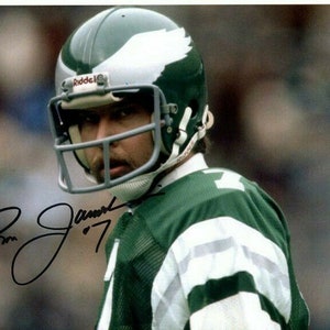 Op de afbeelding: Een American footballspeler die een groen-wit shirt draagt met een witte helm met een groen adelaarlogo. De helm heeft een gezichtsmasker en de woorden "Riddell" aan de zijkant. De speler kijkt naar rechts en heeft een handtekening die "Ron Jaworski '77" zegt.