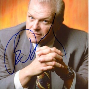 May include: A man in a gray suit with a patterned tie and a watch on his left wrist. He is sitting with his hands clasped in front of him. He has short, gray hair and a mustache. The background is a blurred orange color. The man's signature is visible in the top left corner of the image.