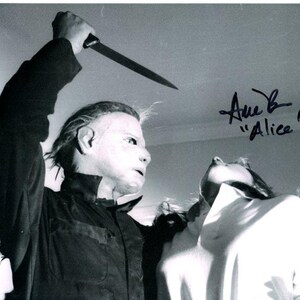 May include: Black and white photograph of a scene from a horror film. A figure in a mask holds a knife above a person in a white shirt. The image is signed "Anne B. 'Alice'".