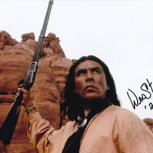 May include: A man with long dark hair holds a black and brown rifle. He is wearing a light brown shirt and a dark scarf. The background is a red rock formation. The image is signed "Wes Studi, 2016".