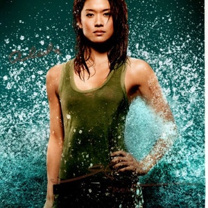 May include: A woman with dark brown hair is standing in front of a blue and green water background. She is wearing a green tank top and a black belt. The water is splashing around her.