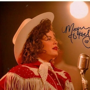May include: A woman wearing a white cowboy hat and a red and white fringed shirt sings into a microphone. The background is a stage with a spotlight. The woman's signature is in the top right corner of the image: "Megan Hilty".