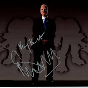 May include: A man in a dark suit and blue tie stands in front of a dark background with two shadowy figures on either side of him. The man is smiling and has a white beard. The figures are holding weapons. The man has signed his name in silver ink on the image.