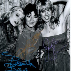 May include: A black and white photograph featuring three women posing together. The image includes signatures in blue, gold, and purple ink. The women have blonde or dark hair and are smiling.